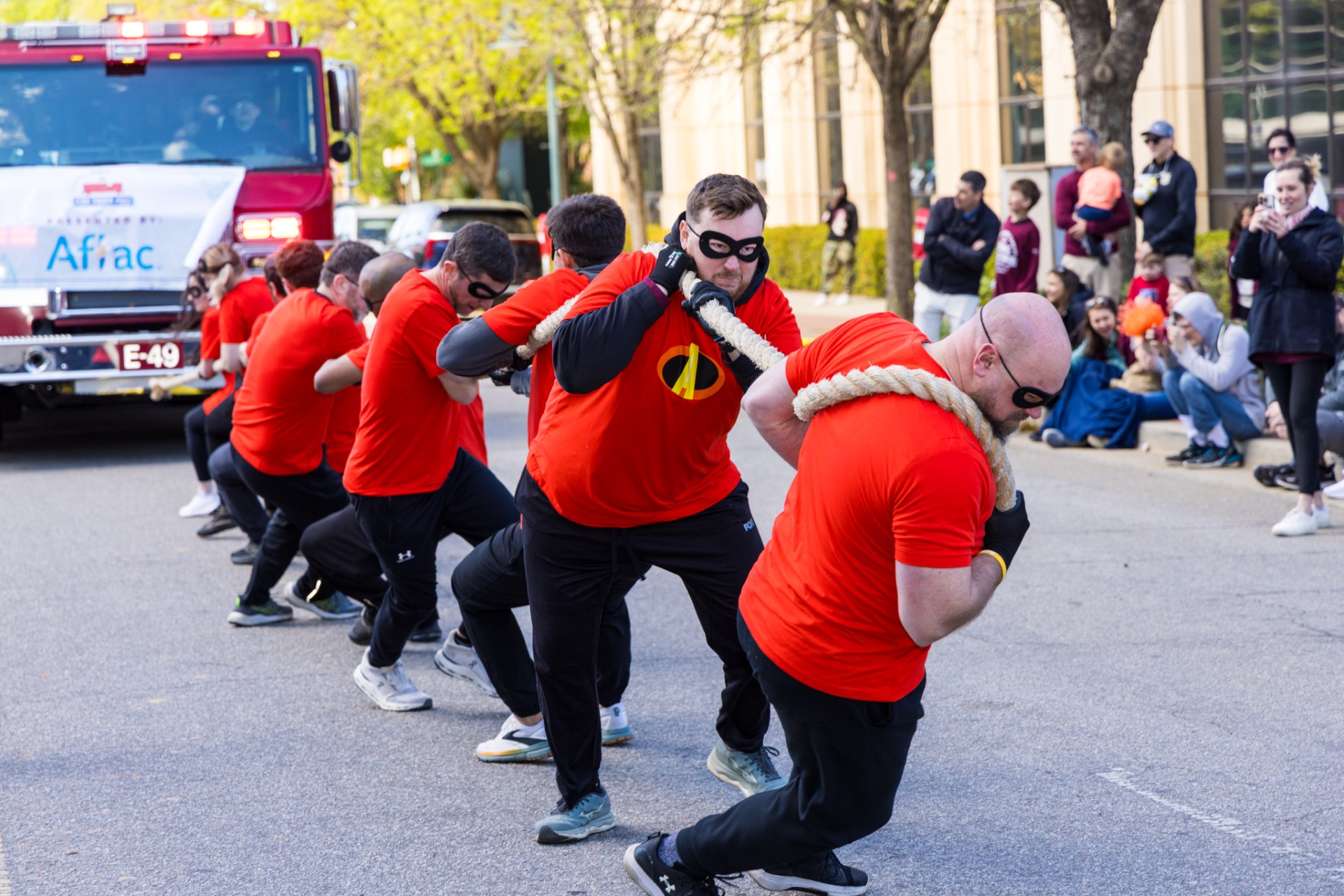 Team of participants in matching red shirts and black masks lean forward in unison, pulling a rope attached to a fire engine during a community fire truck pull event, while spectators watch from the sidewalk.