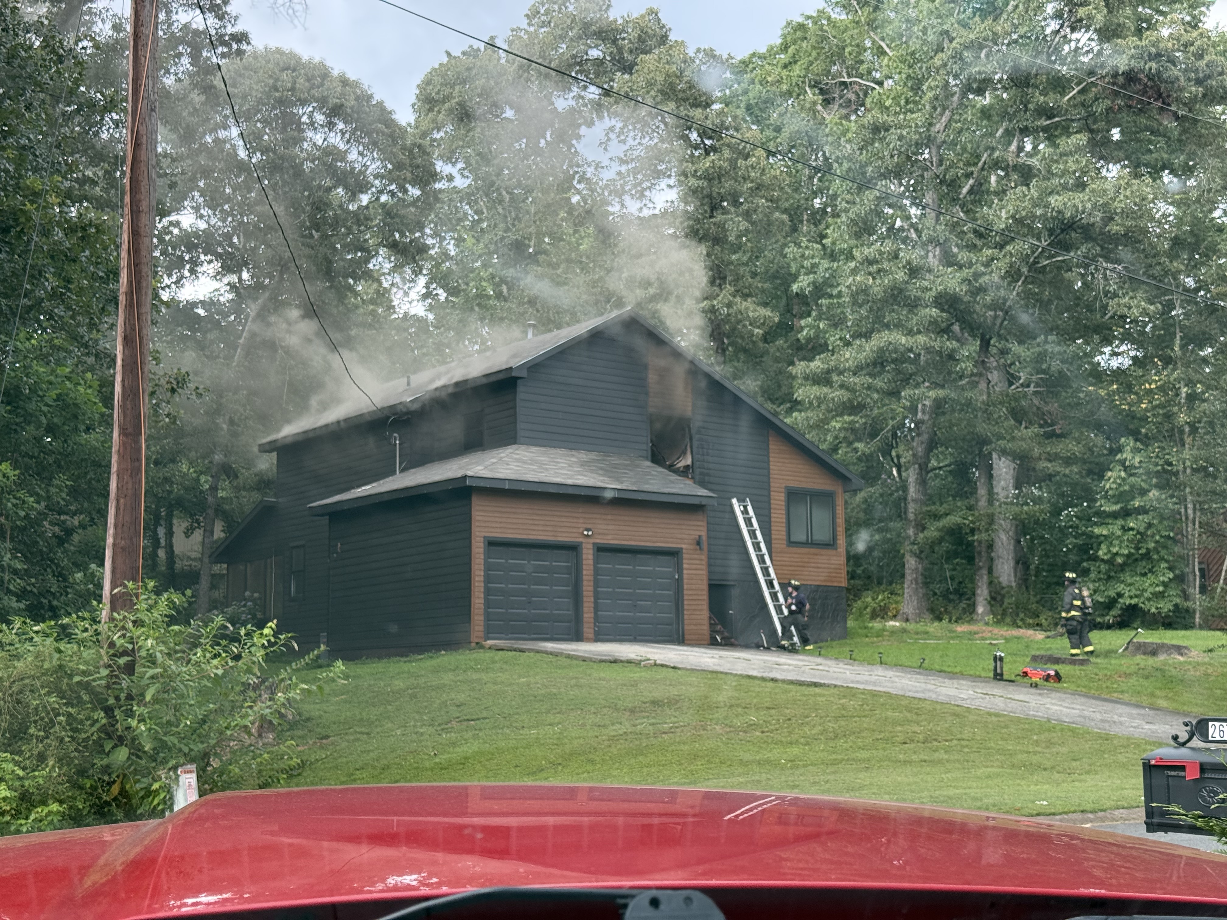 Firefighters work outside a two-story home while light smoke vents from the roof and an upper-story window. A ground ladder is positioned against the house as crews continue overhaul and suppression operations.